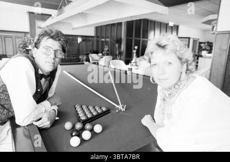 Owners of restaurant Vreeburg, Whizgle News, Dutch Desk, The Netherlands, 1950 - 2000 on 05-1989. These are the elements in the image. The scene captures a moment in a bright, indoor setting that feels both social and relaxed. In the foreground, two individuals lean casually against a green felt pool table, which is adorned with neatly arranged billiard balls, some of which are bright red, others a deep blue, and white. The table is framed by wooden panels that give a warm, classic feel to the space. The man, wearing chunky glasses and a stylish vest over a white shirt, exudes a confident deme Stock Photo