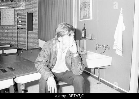 Opening of Lisse school Opening openings inauguration Schools, Whizgle News, Dutch Desk, The Netherlands, 1950 - 2000 on 27-08-1969. These are the elements in the image. In a stark, minimalist setting, a young man is seated on a wooden chair, positioned slightly to the right of the scene. He has a contemplative expression, with his hand resting thoughtfully near his mouth. His hair is styled in a casual manner, and he wears a jacket over a light-colored shirt. The background features a plain wall adorned with a curtain that drapes softly, its color a muted tone that contrasts with the more neu Stock Photo