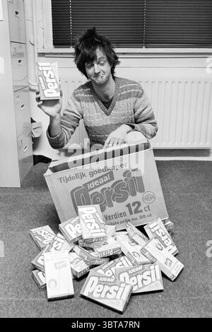 Persil mounting Detergents, Whizgle News, Dutch Desk, The Netherlands, 1950 - 2000 on 19-12-1981. The image shows these topics. The scene features a young man seated on the floor in a brightly lit room, surrounded by products that appear to be laundry detergent boxes. He has unkempt hair, styled in a casual manner, and a playful smile on his face, suggesting a lighthearted mood. His attire consists of a cozy, patterned sweater with shades of gray and brown, which adds to the relaxed atmosphere. In front of him, there’s a large, open cardboard box filled with additional boxes of laundry deterge Stock Photo