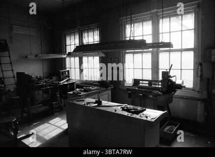 Guided tour Enschede, Whizgle News, Dutch Desk, The Netherlands, 1950 - 2000 on 23-05-1991. The image shows these topics. The scene depicts a workshop bathed in muted, monochromatic tones, primarily shades of gray and black, contributing to a somber ambiance. Natural light filters in through large windows, casting long shadows across the floor and illuminating dust particles that float in the air. To one side, a lathe stands prominently, its metallic surface glistening slightly in the light. Tools are scattered across a central workbench, hinting at recent activity; some tools are placed neatl Stock Photo