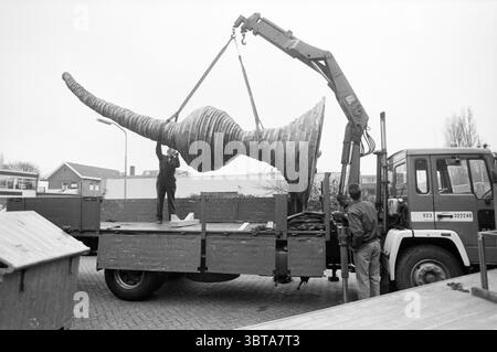 Transport image HD, Whizgle News, Dutch Desk, The Netherlands, 1950 - 2000 on 01-04-1991. The image contains these topics. In a muted monochromatic scene, two individuals are actively engaged in a loading process involving an oversized wooden sculpture. The sculpture has a distinctive spiral shape, reminiscent of a large, ornate horn, with deep grooves and ridges that suggest intricate craftsmanship. The setting is an open area, likely a worksite or fabricating yard, characterized by a mix of concrete and gravel underfoot. There are various vehicles and industrial equipment in the background, Stock Photo