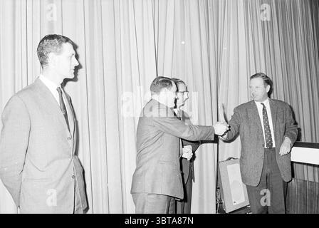 Prizes safety roll call Hoogovens Awards ceremony, Whizgle News, Dutch Desk, The Netherlands, 1950 - 2000 on 01-02-1968. These topics appear in the image. In a formal indoor setting, a group of four men is engaged in a ceremonial moment. The backdrop features thick, draped curtains that create a sense of depth and texture, likely in a muted color that suggests a classic or understated aesthetic. The man on the left stands slightly apart from the others, dressed in a dark suit with a tie, exuding confidence and poise. He gazes toward the action in the center, where two men are shaking hands, in Stock Photo