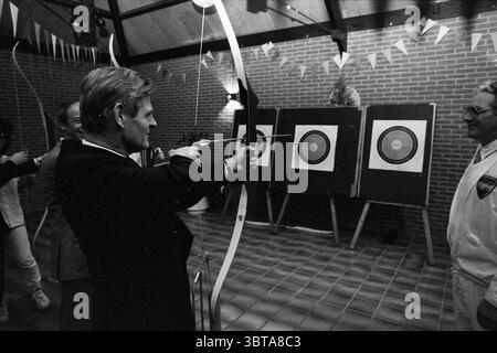 Sports meeting Lisse mayor and alderman Sports and sports week Lisse, Whizgle News, Dutch Desk, The Netherlands, 1950 - 2000 on 18-10-1985. These topics are shown in the image. The scene captures a dynamic indoor archery setting. The room features a high ceiling adorned with wooden beams, creating an inviting yet rustic atmosphere. Brick walls in warm tones provide a contrasting backdrop, and colorful bunting hangs overhead, adding a festive touch to the environment. In the foreground, a person is focused intently on the act of drawing back a bowstring. This individual is dressed in formal att Stock Photo