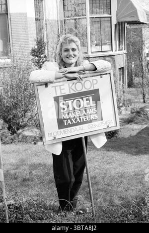 M. Bon the real estate agent, Whizgle News, Dutch Desk, The Netherlands, 1950 - 2000 on 25-03-1993. These are the elements in the image. In a vibrant outdoor setting, a young woman stands smiling confidently in front of a distinctive sign. The scene captures her joyful demeanor as she leans on the sign, which prominently features a frame that suggests its purpose in real estate, giving off a sense of optimism and opportunity. The woman is dressed in a stylish light-colored blazer that contrasts with her bright, spirited expression. Her hair is styled in soft waves, adding to her approachable a Stock Photo