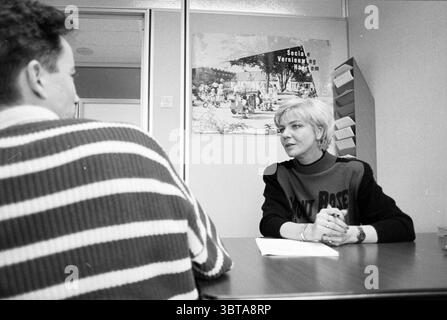 Stella Beek welfare H'lem Haarlem The Netherlands, Whizgle News, Dutch Desk, The Netherlands, 1950 - 2000 on 15-01-1996. The image includes these topics. In a well-lit office environment, a conversation unfolds between two individuals seated at a wooden table. One person, facing away from the viewer, has a casual appearance, wearing a striped sweater with varying shades of white and dark gray. Their hairstyle is short and dark, conveying a relaxed demeanor. Opposite them sits a woman, who is engaging intently in the discussion. She has a stylish short haircut, exhibiting light blonde hair that Stock Photo