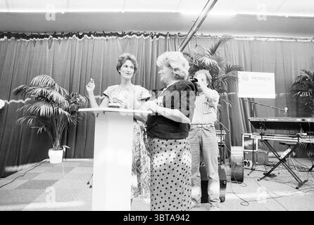 Opening of the Promech company by state secretary. van Rooy, Whizgle News, Dutch Desk, The Netherlands, 1950 - 2000 on 07-09-1989. These are the elements in the image. The scene unfolds in a vibrant indoor setting characterized by a deep red curtain that serves as a backdrop, creating a warm and inviting atmosphere. Two women engage in conversation near a white podium, both exhibiting animated expressions that convey enthusiasm and connection. One woman, dressed in a patterned dress, stands confidently, gesturing with her hand, signaling openness and engagement. The other, wearing a black jack Stock Photo