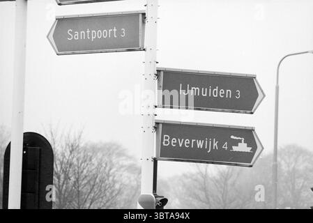 ANWB traffic bollard signs, Whizgle News, Dutch Desk, The Netherlands, 1950 - 2000 on 20-01-1987. These topics are shown in the image. The scene presents a street setting characterized by a foggy atmosphere that creates a soft and muted backdrop. Several directional signs stand prominently, their surfaces displaying a sleek, metallic sheen. The arrows point towards different locations, suggesting a network of routes. Each sign is decorated in a simple yet clean design, featuring a contrasting color scheme of light gray backgrounds with dark lettering. Surrounding the signs, trees appear in the Stock Photo