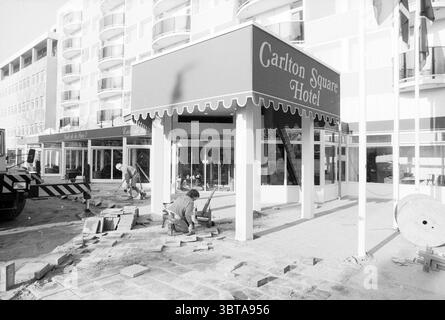 Exterior Carlton Hotel from Maria Stichting Hotels, Whizgle News, Dutch Desk, The Netherlands, 1950 - 2000 on 26-01-1989. These topics are shown in the image. The scene captures an urban construction site in a bustling area. In the foreground, a worker is engaged in laying down gray bricks, meticulously aligning them with care. His clothing is work-appropriate, possibly featuring durable fabrics designed for labor. Surrounding him, scattered tools and materials are present: bricks in various stages of alignment, a wheelbarrow nearby, and a few construction tools like a trowel and a level. The Stock Photo