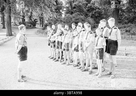 Scouts, Whizgle News, Dutch Desk, The Netherlands, 1950 - 2000 on 07-1963. The image shows these topics. In a sun-drenched park setting, a group of boys stands in a neat line along a gravel path. They wear matching uniforms consisting of light-colored shirts and dark shorts, which are complemented by knee-high socks. Each boy has a distinct scarf tied around his neck, showcasing different colors or patterns, suggesting a playful element of individuality within their uniformity. The leader, positioned a short distance away and facing the group, commands attention. His stance is confident yet co Stock Photo