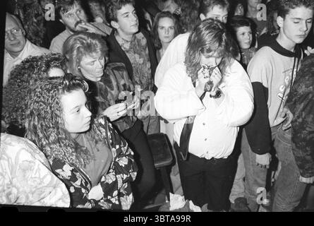 Playback show lantern IJmuiden IJmuiden The Netherlands, Whizgle News, Dutch Desk, The Netherlands, 1950 - 2000 on 25-12-1990. These topics are shown in the image. The scene captures a lively indoor gathering, likely a concert or social event, filled with a diverse crowd. In the foreground, a young woman with voluminous curly hair and a floral-patterned top stands closely to her left, her expression reflective and focused. Next to her, another figure has a neutral expression, holding a handheld camera, suggesting a desire to document the moment. Central to the composition is a man with longer Stock Photo