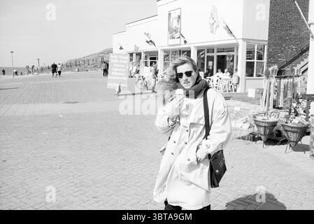 Langevelderslag beach bustle of shops and pedestrians crowds shops and kiosks Noordwijk Langevelderslag, Whizgle News, Dutch Desk, The Netherlands, 1950 - 2000 on 02-04-1989. These are the topics in the image. In a bustling coastal scene, a woman stands prominently in the foreground, wearing sunglasses and a light-colored trench coat, which flows gently in the breeze. She holds a cone of ice cream, and her expression appears playful or carefree. The backdrop features a row of buildings showcasing a mix of storefronts, some adorned with flags that flutter in the wind. The setting is lively, wit Stock Photo