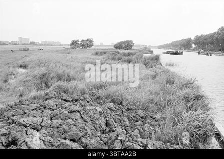 Area behind the Vijfhuizen mill (+ miller) Mills Overviews Vijfhuizen, Whizgle News, Dutch Desk, The Netherlands, 1950 - 2000 on 17-06-1980. The image shows these topics. The scene depicts a serene riverside landscape, characterized by a gentle, muted palette that leans towards shades of gray. In the foreground, a rugged, textured expanse of earth reveals dark, cracked soil, suggesting recent disturbance or erosion. It juts out slightly toward the water, forming a natural boundary between the land and the river. The river itself is calm, with a smooth surface that reflects the overcast sky abo Stock Photo