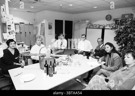 Awards ceremony Primafoon Hoofddorp Hoofddorp The Netherlands, Whizgle News, Dutch Desk, The Netherlands, 1950 - 2000 on 27-12-1995. These are the elements in the image. In a modern office setting, a group of seven individuals is gathered around a large, rectangular table made of light-colored wood. The table is cluttered with various items, including wine bottles, snacks, and paper cups, which suggest a casual gathering or celebration. The room features neutral walls, with a large plant in one corner adding a touch of greenery. The overhead lighting is bright yet soft, creating an inviting at Stock Photo