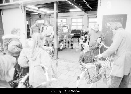 Bicycle inspection for students from the Engelmund school at the police station, Whizgle News, Dutch Desk, The Netherlands, 1950 - 2000 on 30-10-1990. The image shows these topics. In a bustling garage setting, a group of individuals engages in animated conversation and activity. The scene is filled with a mix of warmth and camaraderie, contrasting with the industrial backdrop. People stand both inside and outside the garage, some leaning against bicycles while others are gathered around a man crouched near a bike, possibly inspecting or repairing it. The crouched individual wears a patterned Stock Photo