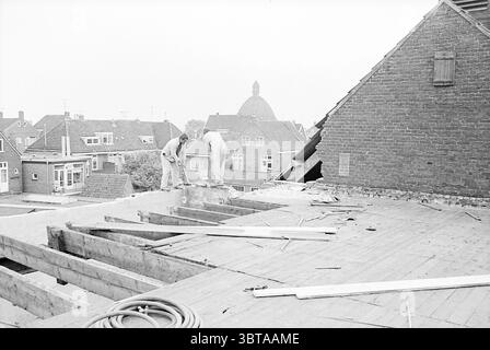 Houses Houtrakpolder Houses and house construction, Whizgle News, Dutch Desk, The Netherlands, 1950 - 2000 on 02-06-1969. The image contains these topics. The scene depicts two individuals engaged in construction work on a rooftop. They appear focused on their task, leaning over a wooden frame that forms part of the structure. Their attire suggests they are well-prepared for the job, with sturdy work pants and shirts, reflecting both practicality and ruggedness. The rooftops in the background create a patchwork of varying hues, primarily shades of gray and brown, reflecting traditional archite Stock Photo