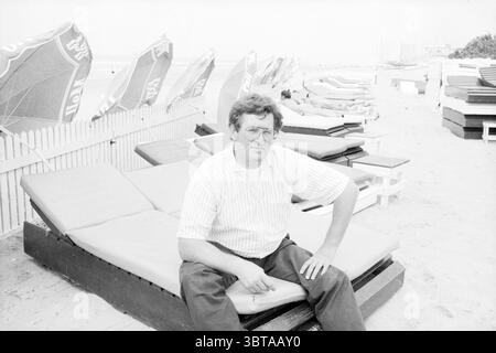 Mr Zonneveld from the Freddy and Paul pavilion Zandvoort Persons Beach and beaches Zandvoort, Whizgle News, Dutch Desk, The Netherlands, 1950 - 2000 on 19-07-1988. These topics appear in the image. In a sunlit beach setting, a man is seated on a wooden lounge near the shoreline. He has a casual demeanor, wearing a light-colored, button-up shirt with thin vertical stripes, paired with dark trousers. The relaxed pose suggests a moment of leisure, as he sits with one leg crossed over the other, leaning slightly forward with a thoughtful expression. The background features several colorful beach u Stock Photo