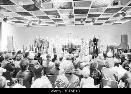 Mary Minstrels in Jansheeren Heemskerk Music, Whizgle News, Dutch Desk, The Netherlands, 1950 - 2000 on 18-02-1978. The image shows these topics. In a spacious indoor venue, a choir stands in formation, evenly arranged in front of a grand piano, which is being played by a seated musician. The choir consists of a diverse group of individuals, dressed in formal attire, predominantly in dark colors that contrast with the light-colored background. Their expressions convey a sense of focus and concentration, as they perform, possibly lost in the music. The setting features a high ceiling with a gri Stock Photo