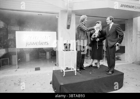 Opening Aronson jeweler Opening openings commissioning, Whizgle News, Dutch Desk, The Netherlands, 1950 - 2000 on 12-11-1986. The image shows these topics. The scene features a celebratory event outside a jewelry store. Three individuals are prominently positioned on a small elevated platform. The first individual, dressed in a stylish, textured blazer, is holding a large glass, likely filled with champagne, while extending it toward the second person, who is standing to their left. This second person, in a dark, elegant dress with a floral arrangement in hand, appears engaged in the toast. Th Stock Photo