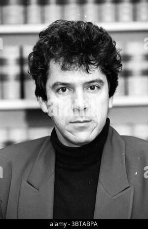 Councilor Houda, Whizgle News, Dutch Desk, The Netherlands, 1950 - 2000 on 04-06-1991. These topics are shown in the image. A man with dark, curly hair stares directly at the viewer, his expression serious and contemplative. He is dressed in a fitted black turtleneck that contrasts sharply with a light gray blazer, giving him a sophisticated yet approachable look. The background is softly blurred, featuring a collection of jars that suggest a setting related to scientific or culinary work. The overall tone is grayscale, lending a classic and timeless quality to the scene. The lighting is soft Stock Photo