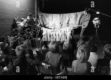 Puppet theater Jan de Noord in the Kadens hall (Harlekinade) Puppets puppet shows puppet shows puppet theater, Whizgle News, Dutch Desk, The Netherlands, 1950 - 2000 on 17-10-1977. These topics appear in the image. In a dimly lit indoor setting, a theatrical performance unfolds before an audience of children. The scene is neatly arranged, with wooden chairs facing a makeshift stage, distinctly illuminated by several bright spotlights positioned on either side. The stage itself is adorned with a rustic backdrop made of fabric and wooden props, suggesting a whimsical environment. A curtain made Stock Photo