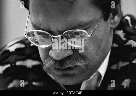 HST overview four-way event Blast Furnace Chess Tournament, Whizgle News, Dutch Desk, The Netherlands, 1950 - 2000 on 12-01-1990. These topics appear in the image. The scene features a man in deep concentration, his face slightly tilted downward, suggesting intense thought or focus. He wears a patterned, dark sweater featuring stripes of varying tones, blending shades of black, grey, and white, which adds texture to his appearance. His eyeglasses are prominent, with round frames that reflect a checkered pattern, possibly from a nearby surface, adding an intriguing element of depth. The man has Stock Photo