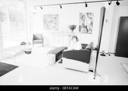 Interior Patrium Heemstede Interior Heemstede The Netherlands, Whizgle News, Dutch Desk, The Netherlands, 1950 - 2000 on 07-09-1989. These are the elements in the image. The scene unfolds in a modern and spacious interior, characterized by minimalist design. A woman sits comfortably on a contemporary dark-colored sofa, her relaxed posture and expression contributing to an inviting atmosphere. She wears a light-colored top that contrasts with the sofa's deep hues. Around her, the room is adorned with carefully selected decor. Two abstract paintings, featuring vibrant splashes of color such as y Stock Photo