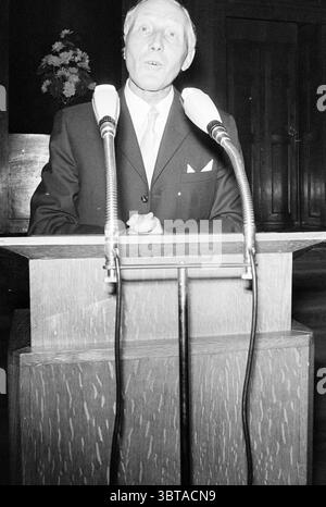 Farewell Alderman Happé Aldermen, Whizgle News, Dutch Desk, The Netherlands, 1950 - 2000 on 07-09-1970. The image shows these topics. The scene depicts a man standing confidently at a podium, delivering a speech or presentation. He is dressed in a well-fitted dark suit with a light-colored tie and a white pocket square, exuding a formal and professional appearance. His face is expressive, suggesting engagement and a strong connection with the audience. The podium is wooden with a polished surface, featuring distinct grain patterns that add warmth to the otherwise formal setting. Two microphone Stock Photo