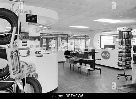 Interior of an Audi and Volkswagen showroom, Whizgle News, Dutch Desk, The Netherlands, 1950 - 2000 on 11-06-1985. These are the elements in the image. The scene depicts a modern service center with a clean and organized layout. The space is predominantly white and light, creating a bright and open atmosphere. To the left, a display of car tires is prominently featured, showcasing several tire models stacked high and draped over a metal rack. In the center, a sleek white counter arcs gracefully, serving as the main service station. Behind the counter, there are multiple service screens display Stock Photo