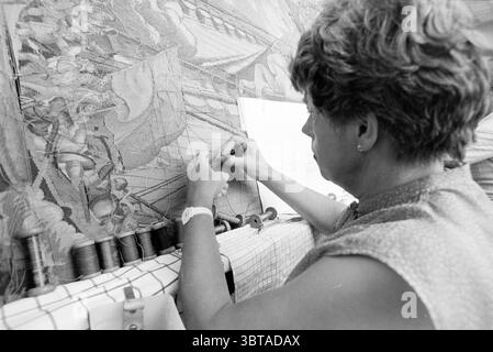 Carpet repair center Leidseplein Haarlem Gobelins tapestries Haarlem Leidseplein The Netherlands, Whizgle News, Dutch Desk, The Netherlands, 1950 - 2000 on 17-07-1985. These topics are shown in the image. The scene captures a skilled artisan engaged in the delicate craft of tapestry weaving. The individual, a woman with short, curly hair, is positioned with her back slightly turned, focusing intently on her work. Her attire, modest and casual, suggests an environment of creativity and dedication. Surrounding her is a rich tapestry being crafted, with intricate designs and vibrant colors of thr Stock Photo