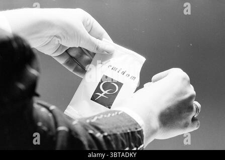 Female condom, Whizgle News, Dutch Desk, The Netherlands, 1950 - 2000 on 11-01-1993. These are the topics in the image. In the scene, two hands are seen holding a small, rectangular package. The hands are well-groomed, with one hand displaying a ring, suggesting a sense of care or attention to detail. The package itself is light in color, primarily white with a hint of shine, hinting at a modern and sleek design. A logo, possibly in black, is visible on the front, featuring an abstract icon that represents femininity. The background is softly blurred and features a gradient effect, transitioni Stock Photo