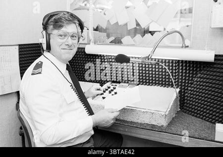 Rob van Rees National Police Driebergen Police Driebergen-Rijsenburg, Whizgle News, Dutch Desk, The Netherlands, 1950 - 2000 on 29-12-1987. These topics appear in the image. The scene captures a man seated in a soundproof recording booth, adorned with a textured wall featuring small black squares that absorb sound. He is dressed in a white shirt with a dark tie, complemented by a badge on his chest, indicating a formal or professional setting. His glasses reflect a glint of light, adding a scholarly touch to his demeanor. A pair of headphones rests atop his head, suggesting he is engaged in au Stock Photo