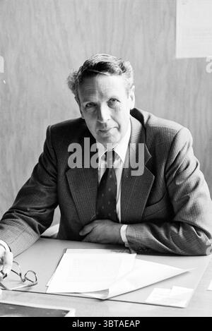 Portrait of Mr Waldorp Portraits, Whizgle News, Dutch Desk, The Netherlands, 1950 - 2000 on 31-10-1979. These topics appear in the image. A distinguished man sits at a simple wooden table, exuding a calm and contemplative presence. He wears a well-tailored suit, characterized by its sharp lapels and a subtle plaid pattern, complemented by a refined tie featuring small, decorative motifs. His hair, slightly tousled, frames his face, accentuating his thoughtful expression. The background is a muted beige or light brown, contributing to an air of professionalism. In the foreground, several sheets Stock Photo