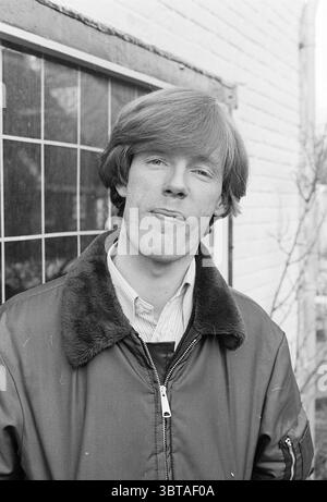 Portrait of a man with a narrow face outside in front of a window with a diamond pattern, Whizgle News, Dutch Desk, The Netherlands, 1950 - 2000 on 20-02-1978. These are the elements in the image. In the scene, a young man stands with a relaxed demeanor, positioned slightly to the side of a grid-patterned window that serves as a backdrop. His hair is a light shade, styled in a voluminous, slightly tousled fashion. He wears a dark, fur-collared jacket, which contrasts with the soft textures of his collared shirt underneath, likely in a lighter hue. The setting has a rustic quality, characterize Stock Photo