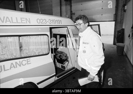 Presentation of new ambulance for Kennemerland, Whizgle News, Dutch Desk, The Netherlands, 1950 - 2000 on 11-1988. The image shows these topics. In a dimly lit garage, the atmosphere feels slightly industrial and functional. A white ambulance, its body marked with vibrant orange stripes, stands prominently. The vehicle's rear door is open, revealing an organized interior that includes medical equipment. A man, dressed in a white jacket adorned with emergency service insignia, steps out beside the ambulance. His hair is slightly disheveled, and there’s an expression of focus on his face as he a Stock Photo