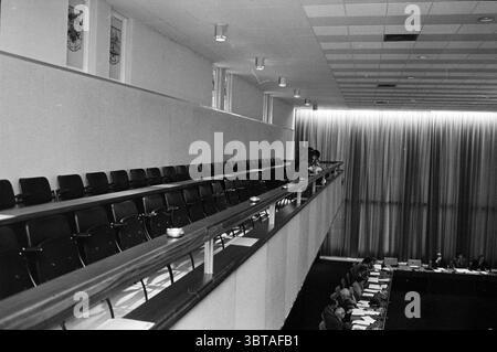 Meeting Social workers Meetings, Whizgle News, Dutch Desk, The Netherlands, 1950 - 2000 on 24-05-1973. These topics appear in the image. The scene depicts a spacious interior, likely a large meeting or conference room. The setting features a high ceiling with a grid of light fixtures illuminating the area softly. The color palette is muted, relying on shades of gray and black, which suggests a serious atmosphere. In the foreground, a long wooden railing separates the upper tier of seating from the lower level. Multiple rows of black chairs are arranged neatly, casting soft shadows on the floor Stock Photo