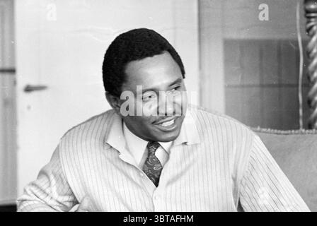 Dr. Janus Anarch Portraits, Whizgle News, Dutch Desk, The Netherlands, 1950 - 2000 on 05-01-1971. The image includes these topics. The scene features a medium close-up of a man with a warm smile, exuding a sense of confidence and friendliness. He has neatly stylized hair, with a slightly curly texture, and his skin tone suggests a healthy complexion. He is dressed in a light-colored, button-up shirt featuring subtle vertical stripes, complemented by a patterned tie that adds a touch of sophistication. The soft lines of his attire contrast with his vibrant expression, embodying a sense of appro Stock Photo
