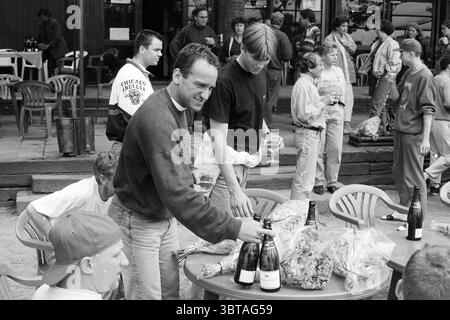 Arrival Kinheim, Whizgle News, Dutch Desk, The Netherlands, 1950 - 2000 on 19-06-1994. These topics appear in the image. In a lively outdoor scene, a group of people is gathered around a circular table. The table, made of a light wood, is laden with multiple bottles of sparkling wine, their glass surfaces reflecting the ambient light. Surrounding the table, people are engaged in conversation, giving off a sense of camaraderie and festivity. The individuals present vary in age and attire. Some wear casual clothes, such as T-shirts and jeans, while others dress more formally. A man standing at t Stock Photo