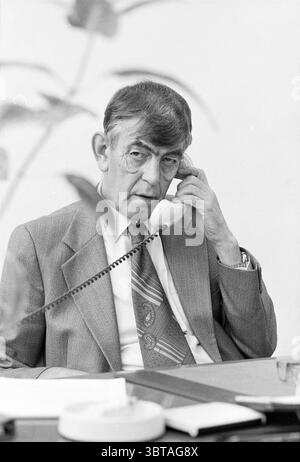 Mr Konst 40 years of PTT PTT postal services city post, Whizgle News, Dutch Desk, The Netherlands, 1950 - 2000 on 25-10-1982. The image includes these topics. In a modest office setting, a man is seated at a desk, engaged in a telephone conversation. He has silver hair, neatly combed back, and his facial expression suggests deep concentration. His attire consists of a light-colored suit and a patterned tie that adds a touch of elegance to his overall look. The desk is simple and functional, featuring a few scattered papers and a solid-colored pen. A round, white object on the desk likely serve Stock Photo