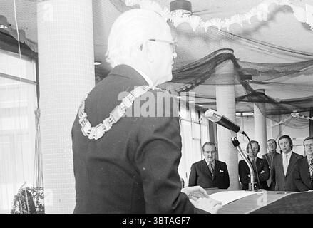 Speech Mayor Beverwijk to billiard players E.K. Speech, Whizgle News, Dutch Desk, The Netherlands, 1950 - 2000 on 11-01-1975. These are the elements in the image. The scene depicts an indoor setting characterized by a formal event or ceremony. A prominent figure stands at a podium, appearing to be delivering a speech. This individual is dressed in a formal black suit, and a distinctive chain of office hangs around their neck, suggesting a position of authority. Their hair is light-colored and styled neatly, indicating a polished presentation. In the background, a group of men is seated or stan Stock Photo