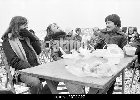 Winter walk Damiate break in the snow, Whizgle News, Dutch Desk, The Netherlands, 1950 - 2000 on 18-02-1979. The image includes these topics. In a snowy outdoor setting, a group of three young people sits around a wooden table. The scene is surrounded by a blur of others in the background, creating a sense of a gathering. The trio at the table is brightly engaged in conversation, their expressions animated; one has thrown their head back in laughter. They are dressed in winter clothing, featuring bulky jackets and scarves, indicating the cold environment. The colored coats contrast sharply aga Stock Photo