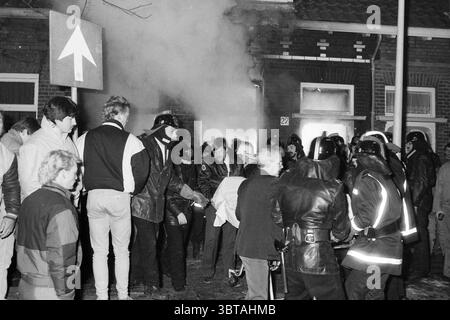 Fire Harmenjansstraat Fires fire brigade Haarlem Harmenjansstraat The Netherlands, Whizgle News, Dutch Desk, The Netherlands, 1950 - 2000 on 11-01-1987. These are the elements in the image. The scene is charged with an intense atmosphere, taking place in a somewhat dimly lit urban environment. Smoke billows from a nearby building, obscuring parts of the view and adding a sense of urgency and chaos. A group of people is gathered closely together, their expressions a mixture of concern and determination. On one side, a cluster of individuals in dark clothing, possibly protestors or bystanders, d Stock Photo
