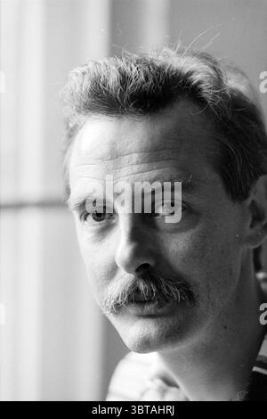 Ruud Scholte Hoofddorp The Netherlands, Whizgle News, Dutch Desk, The Netherlands, 1950 - 2000 on 05-05-1994. These topics appear in the image. In this scene, a man is seated near a window, allowing soft, natural light to illuminate his features. His hair is neatly styled, with a bit of volume and slight wave at the top, conveying a sense of care in his appearance. He has a prominent mustache that adds character to his facial expression. The man gazes thoughtfully, his eyes reflecting both intensity and calm, suggesting a moment of contemplation. The background is softly blurred, highlighting Stock Photo