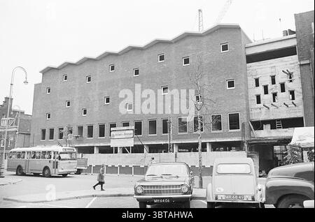 Expansion Post Office. Buildings, Whizgle News, Dutch Desk, The Netherlands, 1950 - 2000 on 07-10-1963. These are the topics in the image. The scene captures a developing urban landscape featuring a multi-story building under construction. The structure's façade showcases a distinct geometric pattern with a series of stepped, wave-like rooflines, creating a visually interesting profile against the skyline. The building’s exterior is primarily composed of red brick, which contrasts with areas of exposed concrete. Large window openings are present, but are not yet installed, giving a glimpse int Stock Photo