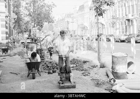 Activities Wilhelminastraat Activities Haarlem Wilhelminastraat The Netherlands, Whizgle News, Dutch Desk, The Netherlands, 1950 - 2000 on 23-09-1986. These are the topics in the image. In a bustling urban environment, a construction scene unfolds along a street lined with charming row houses. The setting is filled with materials typical of a renovation or roadwork project. Two individuals are engaged in labor. One stands at the forefront, operating a heavy machine that vibrates against the surface, indicating they are compacting the ground or paving. They appear focused, wearing practical wor Stock Photo