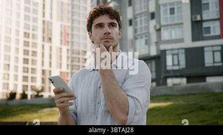 Pensive Caucasian man using mobile phone smartphone thinking thoughtful guy ponder looking around gps navigator maps online male businessman city Stock Photo
