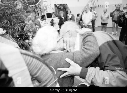 Aunt Toos 80 years old, Whizgle News, Dutch Desk, The Netherlands, 1950 - 2000 on 02-11-1993. These are the elements in the image. The scene captures a warm, heartwarming interaction between two elderly individuals, their faces close together, suggesting a moment of joy and connection. The woman has short, white hair, adorned with glasses, and is dressed in a cozy, textured sweater. The man, also gray-haired, leans in with a soft smile, offering a sense of affection. Surrounding them are numerous people, blurred in the background, indicating a lively gathering. Some participants appear to be e Stock Photo