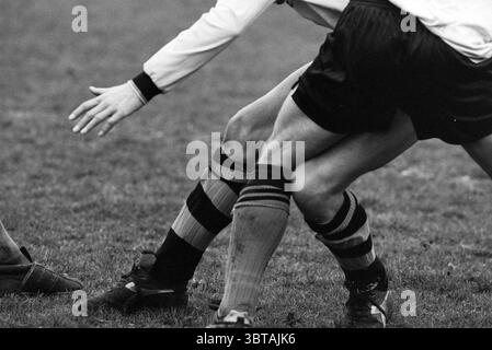 Football Scots - HBC, Whizgle News, Dutch Desk, The Netherlands, 1950 - 2000 on 21-03-1992. These are the topics in the image. The scene captures dynamic movement on a grassy field, where two players engage in an intense moment of competition. One player, clad in a white long-sleeved shirt, reaches out with a hand, showing determination and focus. His black shorts contrast with bold vertical stripes on his socks—orange and black—which draw the eye and emphasize the action. The other player is partially visible, showcasing strong legs in motion as he shifts to defend. His black shorts blend wit Stock Photo
