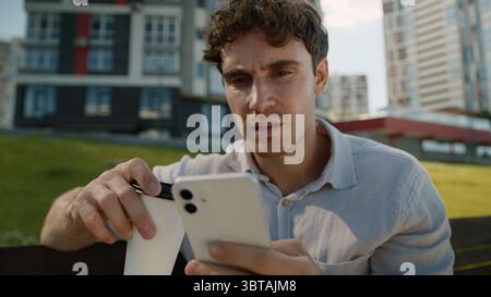 Shocked dissatisfied Caucasian young man holding cup of coffee user mobile phone smartphone male city amazed guy bad news message online student Stock Photo