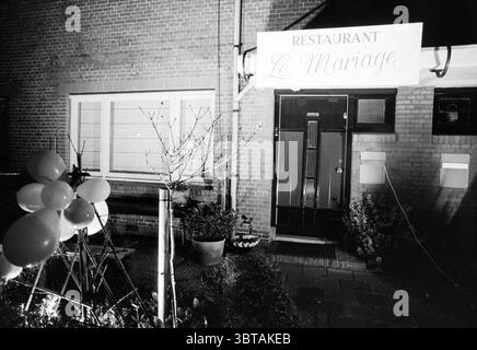 Elk's family married for 12 1/2 years, Whizgle News, Dutch Desk, The Netherlands, 1950 - 2000 on 21-01-1995. These are the topics in the image. The scene captures a nighttime exterior of a quaint establishment, exuding a charming yet understated vibe. The building showcases classic brickwork, with a two-tone design that features both a warm reddish-brown and soft beige, creating a cozy atmosphere. To one side, a tall, elegant entrance door stands out with its sleek, black finish and art deco detailing, hinting at a stylish interior. Large windows flank the door, their muted light softly illumi Stock Photo
