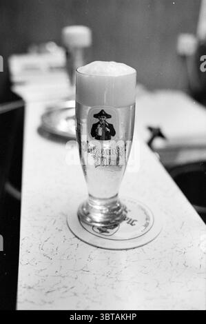 Etc. etc reportage Germany Reportage Tourism tourists, Whizgle News, Dutch Desk, The Netherlands, 1950 - 2000 on 09-09-1980. The image shows these topics. The scene centers around a tall, slender glass filled with a pale golden beer, topped with a thick, frothy head that cascades slightly over the rim. The glass has an intricate logo featuring a pirate, which adds a whimsical touch to the overall composition. The background is softly blurred, with hints of warm wood tones and muted colors that suggest a cozy, intimate setting. A white surface with a subtle texture complements the scene, provid Stock Photo